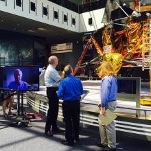 Three speakers with backs to camera look at the lunar module. Next to them is a TV screen with guest astronaut. 