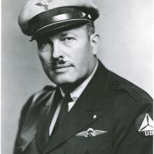 Half length black and white photograph of man, wearing a full-brim light-colored wheel hat with winged insignia and uniform with "RT" insignia wings over breast pocket. He has a mustache with pointed tips.