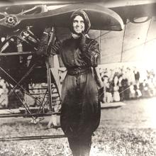 Woman in flying gear poses in front of an airplane. Right hand on propeller, left hand on her chin. Crowd in background.