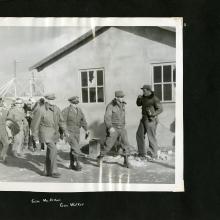 Black scrapbook page. Centered photo in photo corners: 3 men in uniform (Gen. Douglas MacArthur 3rd on the left) walk in front of a building. Background left: group in uniform following. Background right: Man in leather jacket and cap.