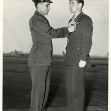 (Left) Man in military uniform and hat pins an award on man in cap on right. Air field with aircraft in background.