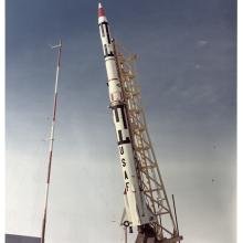 White rocket with red and black details, USAF painted on side, on yellow scaffolding.  Car and red and white pole in the background