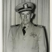 Half-length portrait of man in light colored summer uniform with hat, posed standing in front of a light-colored curtain