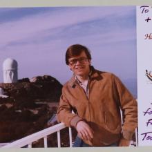 (left) Man with brown hair and glasses, wearing a tan jacket, stands at a railing. Background is an observatory with white buildings on a hill. (right) preprinted message "Happy Holidays" with drawing of horses and sleight. Handwritten note.