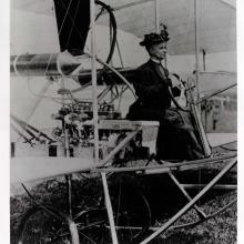 Woman seating at the steering wheel of an early airplane, wearing a hat