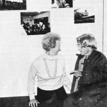 Black and white photograph of two women in front of a gallery wall with four photographs. Woman on the left wears a white shirt and a necklace. Woman on the right wears a dark jacket.