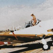 Right side view of woman in hat and khaki shorts stands in the cockpit of a white aircraft with yellow wings and red nose and tail. Front wheels are down.