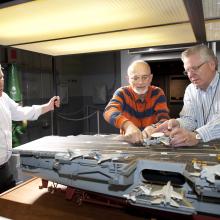 curator Chris Moore (left) looks on as Steve Henninger (center), assisted by Paul Moore, frees the E-2C on the model of the USS Enterprise.