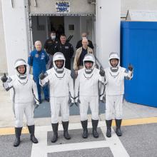 The crew of SpaceX Crew-1 stands in white spacesuits outside of the Neil A. Armstrong Operations and Checkout Building