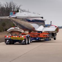 The Douglas DC-3 is on a truck on a highway. It is moving away from the camera. 