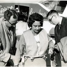 Black and White photograph: Airplane in the back has writing: Spirit of Columbus. Woman in the center has dark hair and wears and inflatable vest. Men on left and right wear glasses and are turning towards the woman.