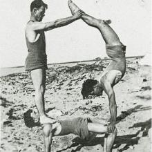 Doolittle and friends do tricks on the beach. Doolittle on the bottom holds up his other two friends who create a pyramid. 