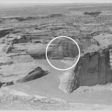 A black and white photograph showing large vertical rock formations that house cliff dwellings. The dwelling area is circled by a superimposed white circle.