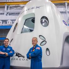 two astronauts in blue flight suits in from of capsule