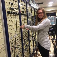 Woman stands in front of a large control board with multiple wires and access points. 