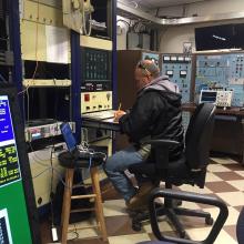 Man works at a small desk surrounded by equipment. 