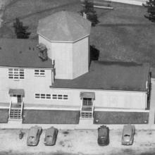 Aerial photo of a building with one large silo and cars parked out front. 