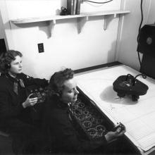 Two women sit in front of controls and plotting table. 