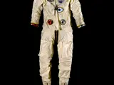 A cream colored space suit with accents of red and blue posed against a black background. There are boots attached but no helmet or gloves.