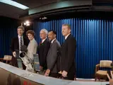 A group of 5 people stand shoulder to shoulder in front of a model of a telescope behind a table. On the other side of the table are members of the press.