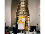 A large golden colored cube inside a metal conical encasing is surrounded by various technicians performing different activities on the structure.