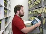 A man opens an archival box and holds a news magazine. On the magazine is a picture of Earth from space. Behind him are shelves of archival boxes. 