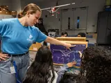 A young woman in a camp t-shirt points to a point on a board with a model of an airplane, as campers work on the plane. 