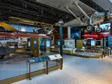 A Museum gallery full of 1920s and 1930s airplanes. Hanging from the roof at the entrance is a silver airplane with a propeller. Text on the side of the plane read "Spirit of St. Louis." Behind it, more aircraft is evident, including a biplane and a small red airplane. There are also metal cases to hold objects. 