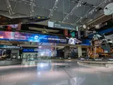 A museum gallery showing highlights from the Museum's collection, including an experimental black plane, a lunar module, satellites, and an electronic sign welcoming visitors the museum. 
