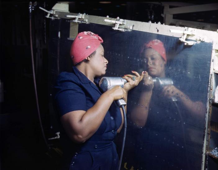 A woman in a red kerchief drives rivets into a silver reflective surface. 