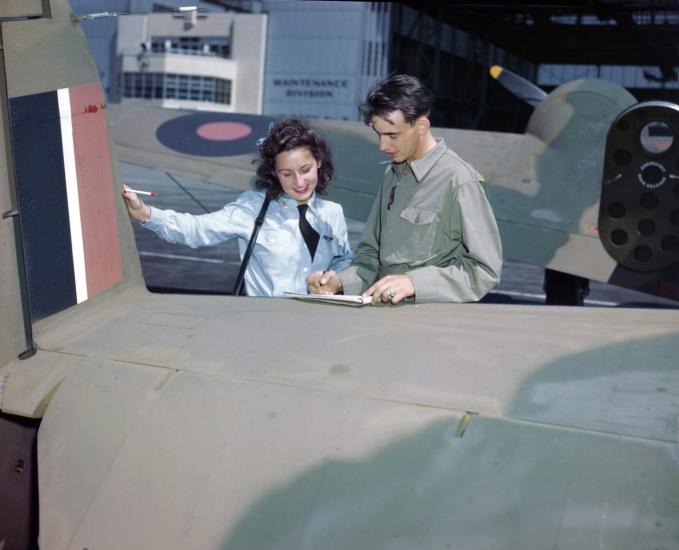 A woman and a man look at a document, which appears to be balanced on an airplane. 