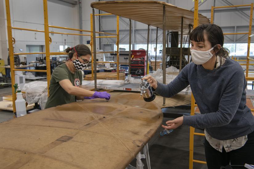 Two conservators, wearing masks, work on the Wright Flyer. 