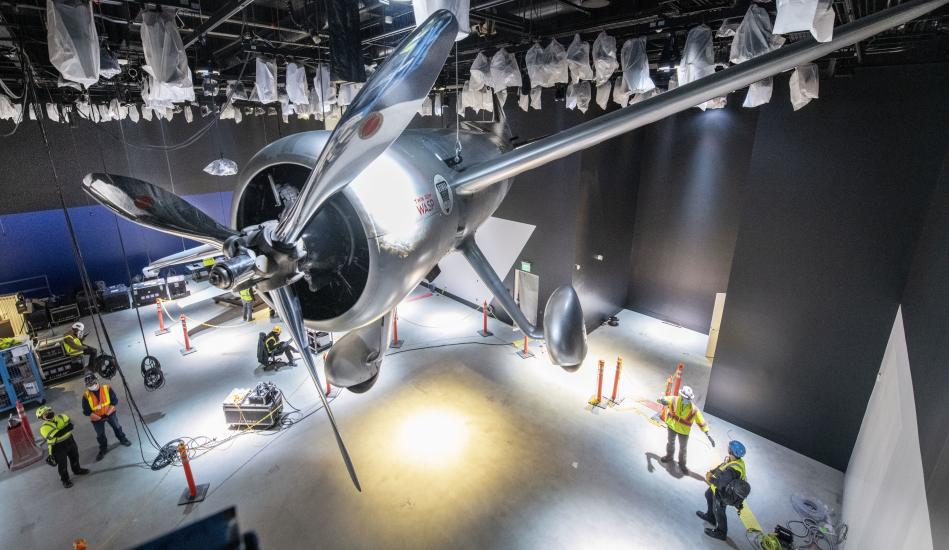 Aircraft hanging in under construction gallery with people in yellow construction vests in the background