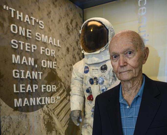 Man in blue shirt stands in front of exhibit case with spacesuit inside.