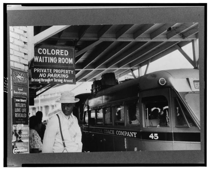 A Black man stands next to a bus and a sign that reads "Colored Waiting Room"