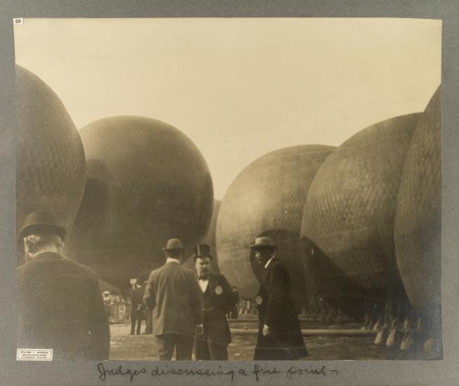 Black and white photo. In the center are three men dressed in suits, the two in the right wearing derbys, the middle man in a top hat. Five hot air balloons in the background. The back of a man in a suit and derby is in the foreground in the lower left corner.