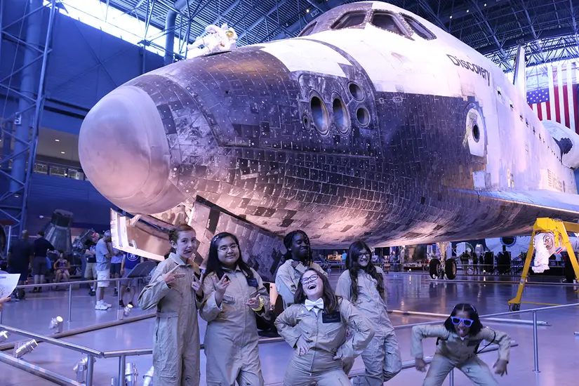 Young women pose in silly positions in front of the Space Shuttle Discovery. 