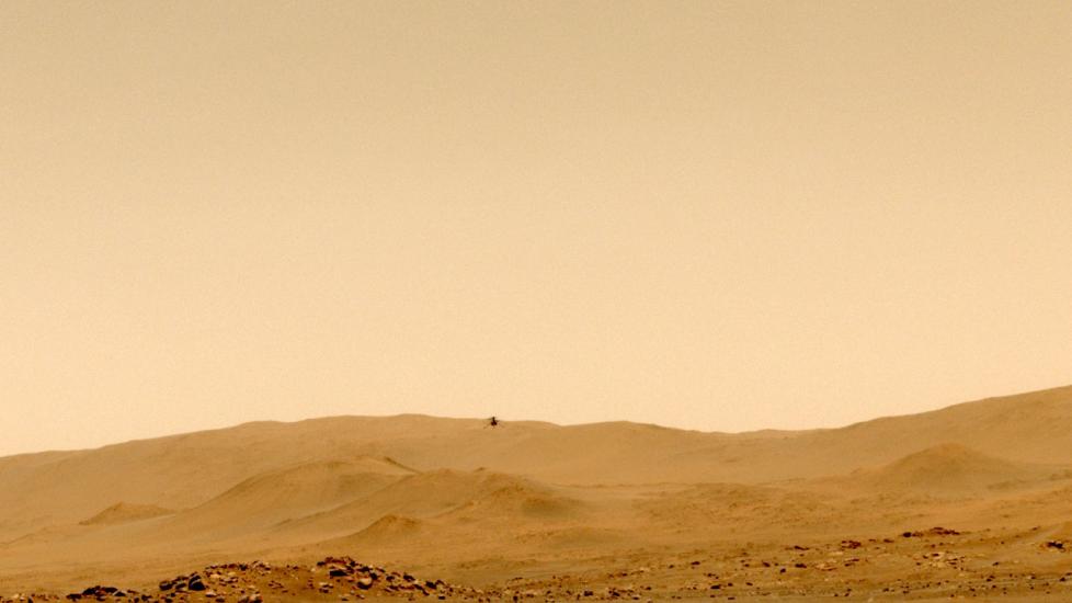 A view of the orange surface of Mars with hills in the background and a barely visible device flying above in the air.