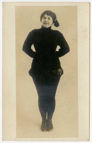 A woman in a jaunty hat stands with her hands behind her back. 