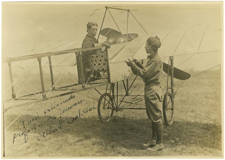 Black and white photograph. One-half right rear view of a man sitting in the cockpit of a monoplane, wearing a helmet with goggles pushed up. To his right a man stands on the ground (back mostly turned) with his hands up in the air. He is wearing a cap.