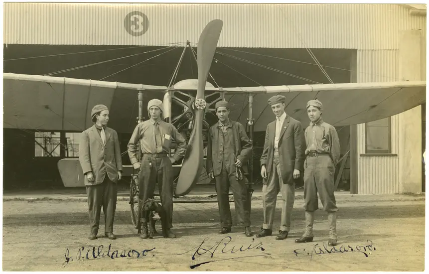 Five men stand in front of a monoplane with a two blade propeller. Background: a hanger made of corrugated metal and to the left of center is the number 3 inside a circle.  From left to right: first man in a suit jacket facing slightly to the right with a backwards cap. second man is wearing a backwards cap and does not wear a jacket and has his left hand in his pocket. The third man is on the other side of the propeller is wearing a cap and unbuttoned jacket.  The fifth man stands with his hands behind him