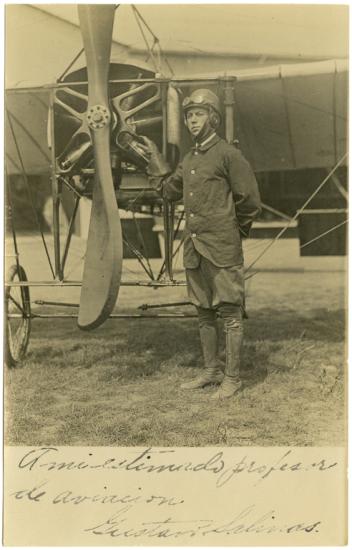 Man stands to the right of a two-bladed propeller with the blades running from the sky to the ground. He is wearing a leather cap with goggles pushed up over the cap and a button down jacket.