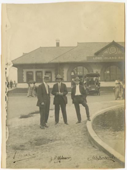 Black and white photo. Three men in suits in foreground. Man on left, turned slightly to his right side is turned to his right and wears a flat brimmed hat.  Man in center wears striped tie and fedora and holds his hands together. Man on right has his left foot up on the edge of a found, wearing a fedora and pale tie. Behind them to the left are two women, one with a parasol. Behind to the right is the back of a car. In background is a building with three rectangular windows on the left and arched windows.