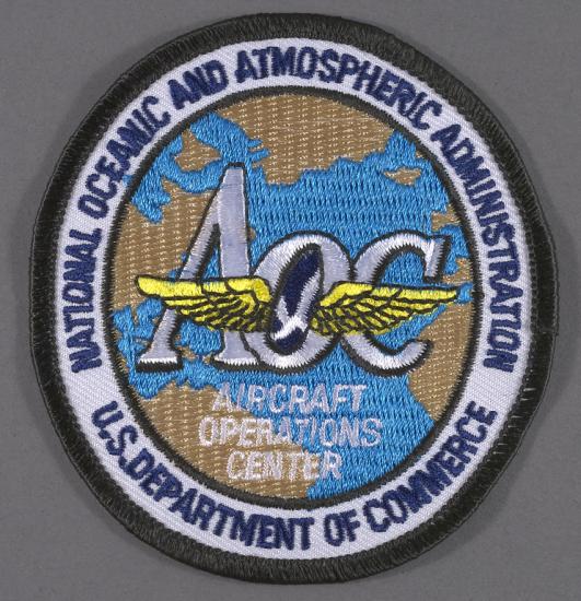 Embroidered National Oceanic and Atmospheric Administration (NOAA) Aircraft Operations Center (AOC) insignia; gold wings in center over white letter text "AOC", "AIRCRAFT OPERATIONS CENTER"; background map of the Atlantic Ocean depicted in blue with tan continental land masses; map surrounded by white band with navy blue letter text "NATIONAL OCEANIC AND ATMOSPHERIC ADMINISTRATION U.S. DEPARTMENT OF COMMERCE"; black edge.