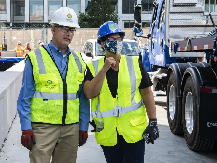 Two people stand with safety vest and hardhats on outside.