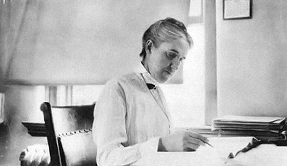 Black and white image of Henrietta Swan Leavitt sitting at a desk and photographed from a side angle. She is writing with a pen in her hand.