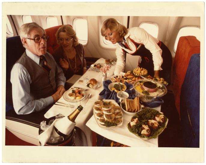 A color photo of a flight attendant serving food to two passengers aboard a plane.