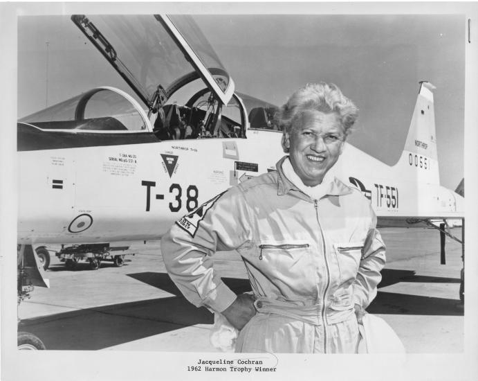 An aged Jackie Cochran posing with her hands on her hips and smiling and a T-38 Talon aircraft in the background.