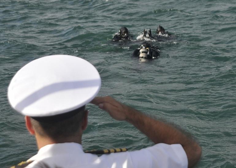 A man in a white military uniform and hat is pictured from behind as he salutes divers in a body of water.