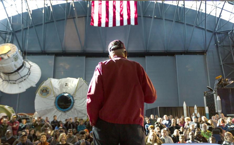 Man with red jacket and cap stands on stage with back towards camera, audience slightly below